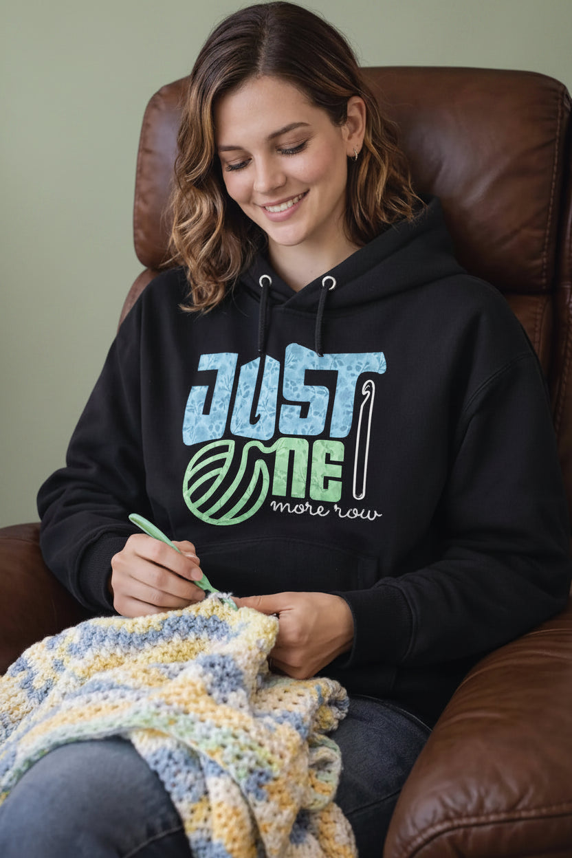 Woman wearing a black oversized hoodie with 'Just Me More Yarn' text, sitting on a brown leather chair and knitting.