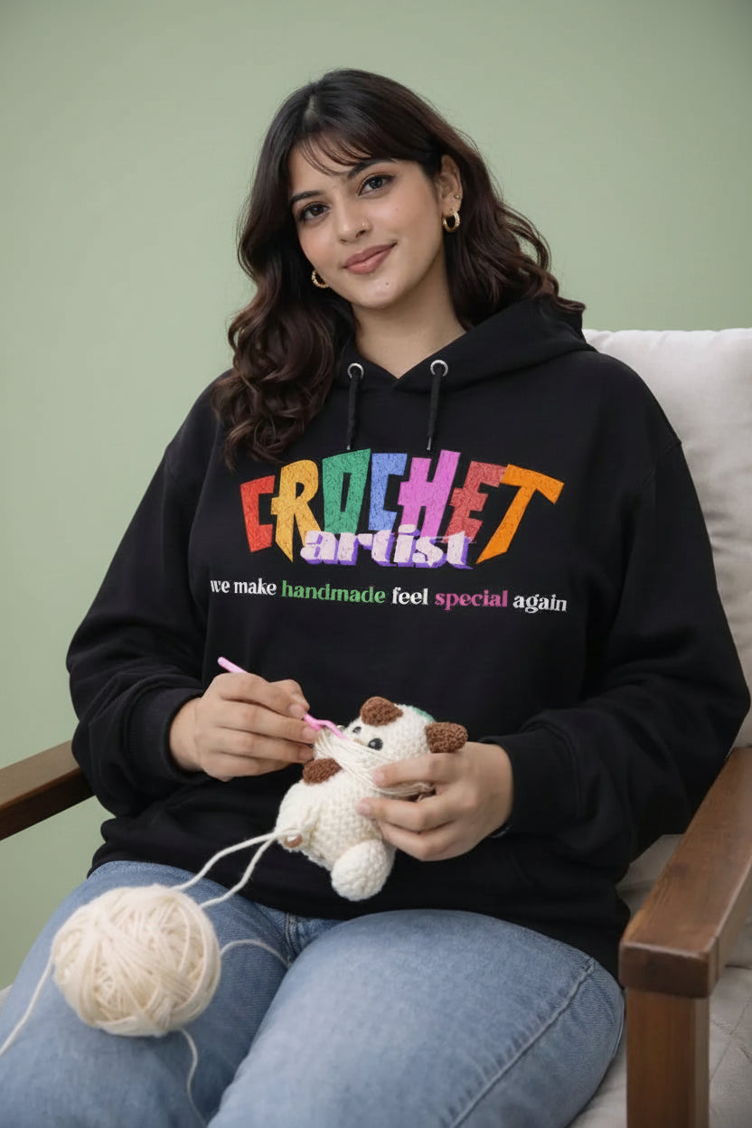 Woman wearing a black hoodie with 'Crochet Artist' text, sitting on a wooden deck with mountains 