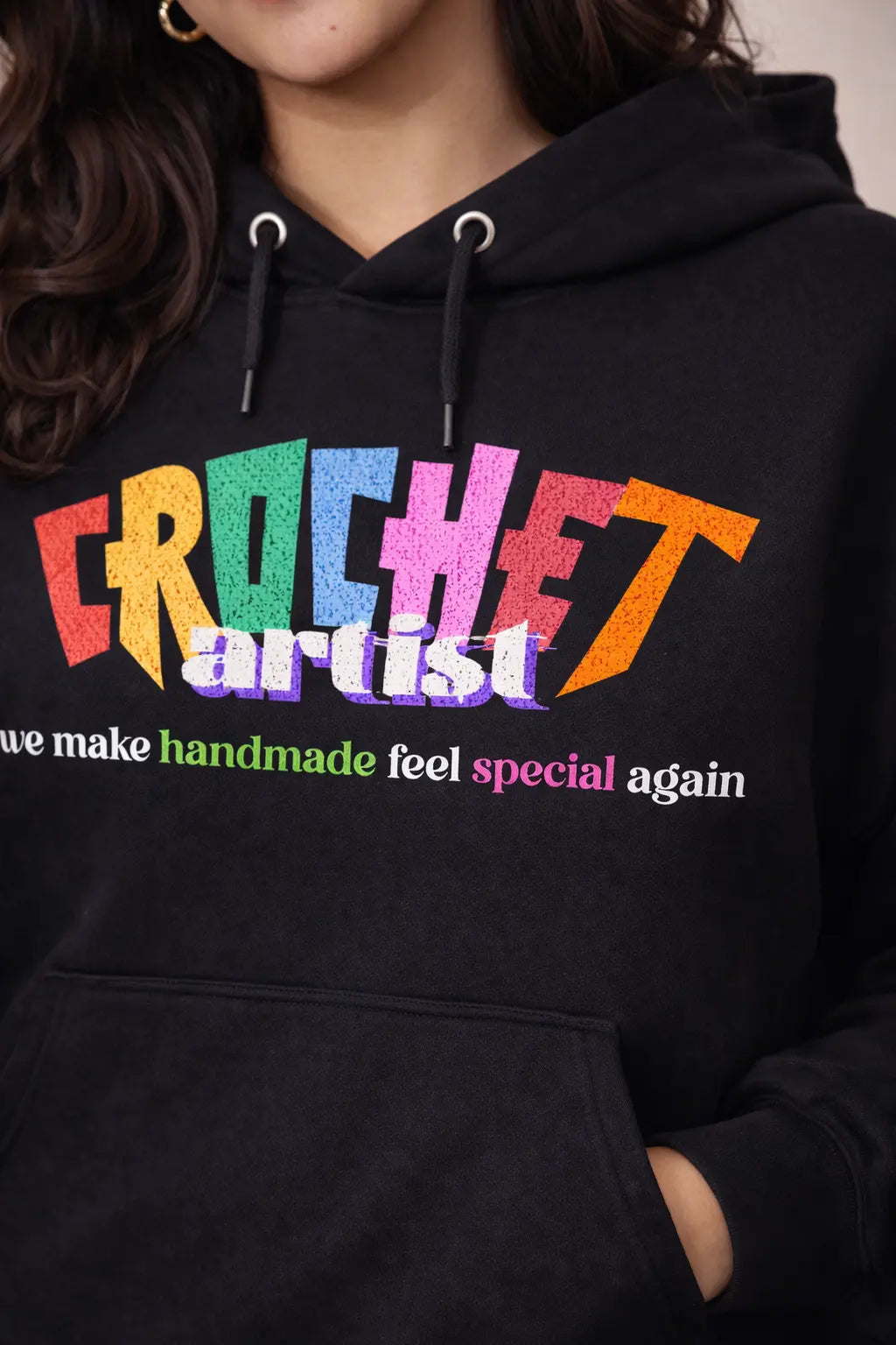 Black hoodie with colorful text 'Crochet artist makes handmade feel special again' on a neutral background